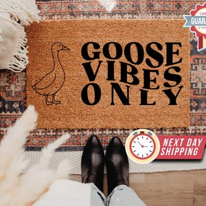May include: A rectangular coir doormat with a goose illustration and the text "GOOSE VIBES ONLY". The mat is a natural brown colour. A guarantee badge is visible in the top right corner. The image also shows a person's feet.