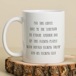 May include: A white ceramic coffee mug with a curved handle. The mug features black text that reads, "MAY THIS COFFEE GIVE ME THE STRENGTH TO ENDURE ANOTHER DAY ON THIS FUCKING PLANET WITH DONALD FUCKING TRUMP AND HIS FUCKING CULT."