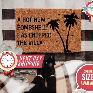 May include: A rectangular brown coir doormat featuring black text that reads A HOT NEW BOMBSHELL HAS ENTERED THE VILLA. The design includes a black silhouette of two palm trees and several small birds flying in the background on the right side.