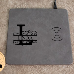 May include: A grey leather wireless charging pad with a black monogrammed initial "L" and the name "LINDA" in a split monogram style. The charging pad has a wireless charging symbol on the right side.