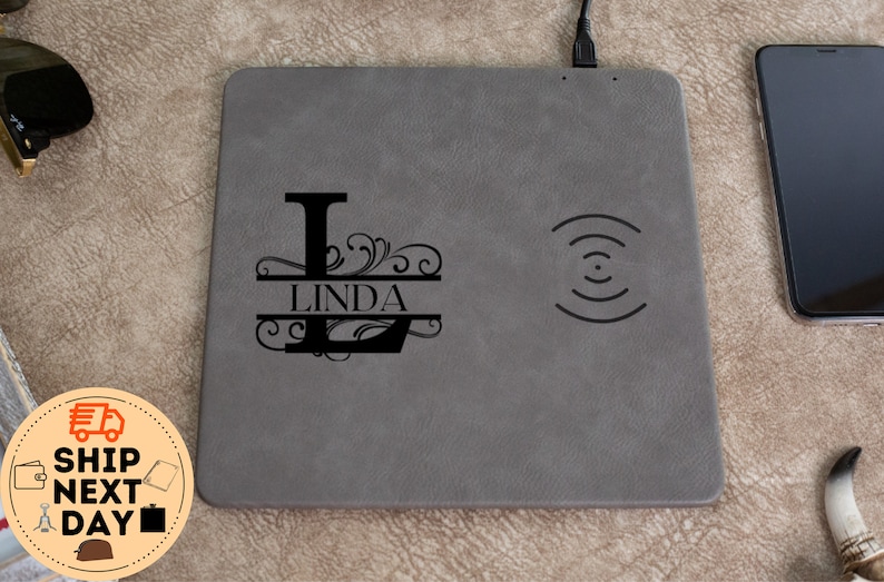 May include: A grey leather wireless charging pad with a black monogrammed initial "L" and the name "LINDA" in a split monogram style. The charging pad has a wireless charging symbol on the right side.