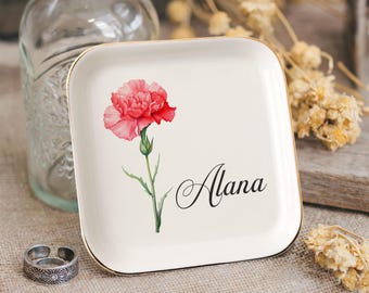 May include: A square, cream-coloured trinket dish with a gold rim, featuring a watercolour carnation and the name "Alana" in elegant script. A silver ring sits beside the dish. A clear glass bottle is in the background.