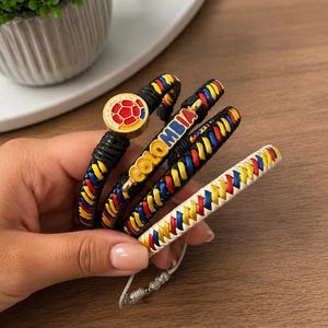 Könnte beinhalten: Ein Set aus vier geflochtenen Armbändern mit gelben, blauen und roten Lederbändern. Ein Armband enthält ein kreisförmiges Emblem mit einem rot-gelben Fußballmotiv, während ein anderes den goldfarbenen Schriftzug COLOMBIA in Blockbuchstaben zeigt.