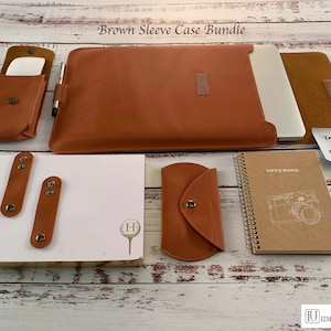 Puede incluir: Un conjunto de fundas de cuero marrón para portátiles, cuadernos y otros dispositivos electrónicos. El conjunto incluye una funda para portátil, un cuaderno, un estuche para bolígrafos, una funda para teléfono y dos organizadores de cables. La funda para portátil tiene una cremallera y una correa de cuero marrón. El cuaderno tiene una cubierta marrón con un diseño de cámara blanca y las palabras "Notebook" impresas en la parte delantera. El estuche para bolígrafos tiene una solapa de cuero marrón con un cierre de botón. La funda para teléfono tiene una solapa de cuero marrón con un cierre de botón. Los organizadores de cables son correas de cuero marrón con broches metálicos. El conjunto se muestra sobre una superficie de madera.