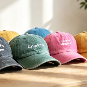 Pode incluir: Uma coleção de seis bonés de beisebol em várias cores, incluindo cinza escuro, amarelo, azul, verde e rosa. Cada boné apresenta texto bordado em branco, com frases como Your Child's Name, Charlotte e Favorite Daughter visíveis na frente.