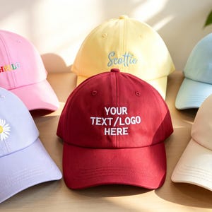Peut inclure: Un ensemble de six casquettes de baseball aux couleurs pastel et unies, notamment rose, jaune, bleu clair, lavande, bordeaux et crème. Chaque casquette présente une broderie unique, incluant le texte HELLO, Scottie, MR, Bride, une marguerite et YOUR TEXT/LOGO HERE.