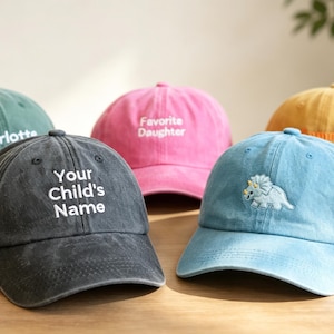 Könnte beinhalten: Fünf Baseballkappen in verschiedenen Farben, darunter Grün, Anthrazit, Pink, Blau und Gelb. Die anthrazitfarbene Kappe trägt den Text 'Your Child's Name', die grüne 'Charlotte', die pinke 'Favorite Daughter' und die blaue zeigt einen Dinosaurier.