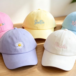 May include: Five baseball caps in pastel colours including pink, yellow, light blue, lavender, and white. The pink cap features the word HELLO, the yellow cap says Scottie, the light blue cap has the letters MK, the lavender cap has a daisy, and the white cap says Bride.