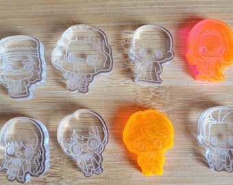 May include: Eight clear acrylic charms featuring cartoon characters. The charms are in the shape of people with different hairstyles and clothing. One charm is orange.