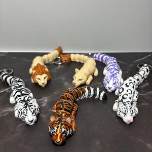 May include: A collection of animal-shaped keychains. The keychains feature various big cat designs, including tigers, leopards, and lions. The keychains are in a variety of colors, including black and white, orange and black, and purple and white.