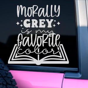May include: White vinyl decal with the text "Morally Grey is my Favorite Color" in a whimsical font. The text is arranged over an open book illustration, suitable for car windows or laptops.
