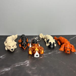 May include: A collection of dog-shaped keychains in various breeds and colors. The keychains include a cream-colored dog, a black and orange Doberman, a beagle, a pug, and an orange spaniel. Each keychain is lying down.