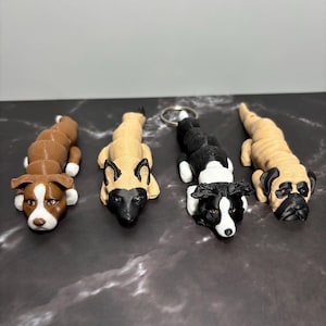 May include: Four dog-shaped keychains in various breeds, including a brown and white pit bull, a tan and black dog, a black and white border collie, and a tan pug. Each keychain is lying down, with detailed features.