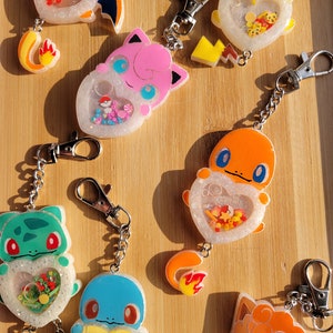 May include: A set of eight colorful Pokemon character keychains. Each keychain features a different Pokemon character with a heart-shaped charm filled with glitter and colorful beads. The keychains are attached to silver chains.