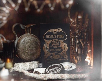 May include: A black book with gold lettering titled "The Devil's Tome" by Shiva Honey, surrounded by various occult and spiritual items, including a silver pitcher, a white sage smudge stick, and a beaded necklace.