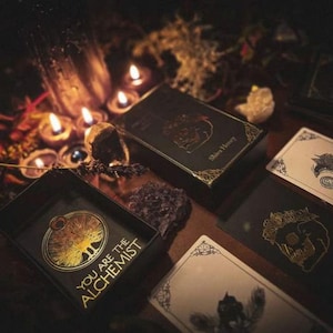 May include: A dark, moody image featuring tarot cards and boxes with gold foil accents. The text "YOU ARE THE ALCHEMIST" is visible on one box. Lit candles and crystals add to the mystical aesthetic. The overall theme is divination and spirituality.