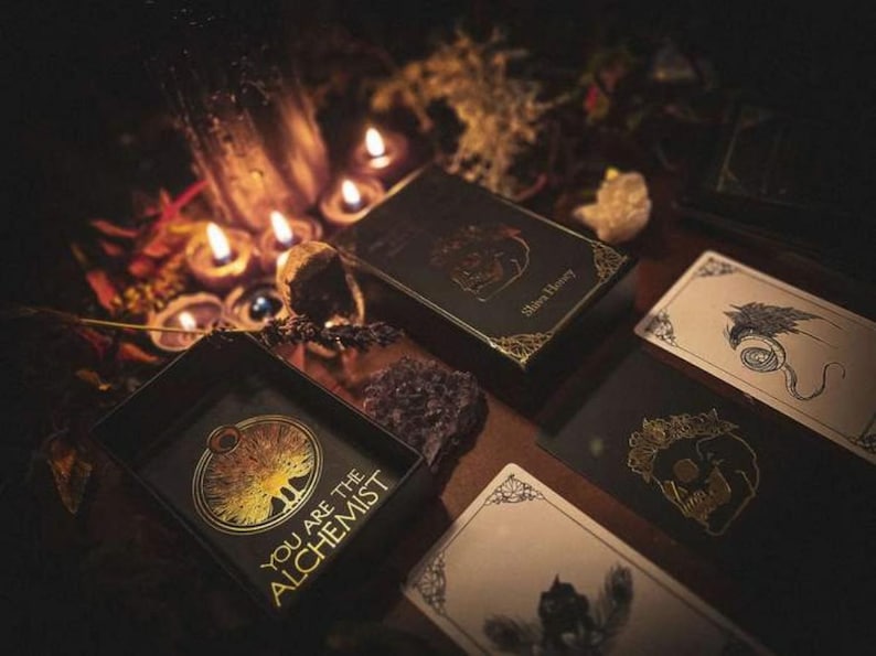 May include: A dark, moody image featuring tarot cards and boxes with gold foil accents. The text "YOU ARE THE ALCHEMIST" is visible on one box. Lit candles and crystals add to the mystical aesthetic. The overall theme is divination and spirituality.