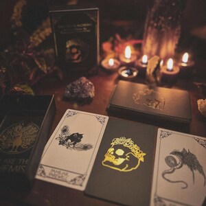 May include: A tarot card deck with a skull design, open box, and several cards are displayed on a wooden table. The cards feature illustrations of a cat, skull, and eye. Lit candles and crystals are in the background.