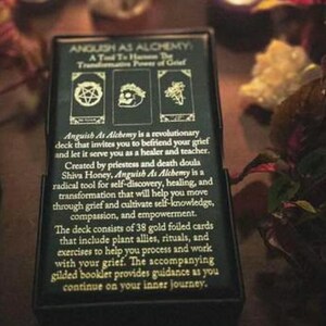 May include: A black box with the text "Anguish As Alchemy" and a description of the deck's purpose. The box features three small images and text describing the deck's use for self-discovery, healing, and transformation.