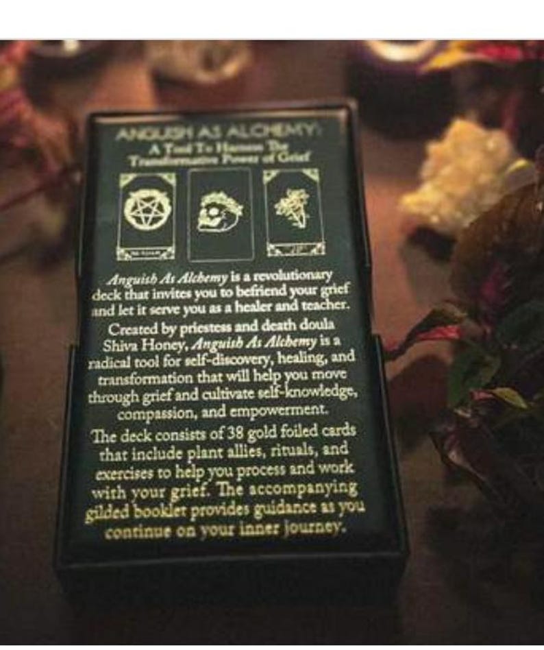 May include: A black box with the text "Anguish As Alchemy" and a description of the deck's purpose. The box features three small images and text describing the deck's use for self-discovery, healing, and transformation.