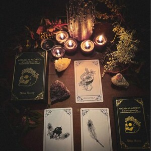 May include: A dimly lit scene featuring tarot cards, crystals, and lit candles. Two decks of cards with gold accents and the text "Anguish as Alchemy" are visible. Several tarot cards are displayed, along with a large candle and smaller tea lights.