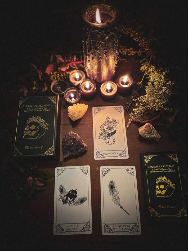 May include: A dimly lit scene featuring tarot cards, crystals, and lit candles. Two decks of cards with gold accents and the text "Anguish as Alchemy" are visible. Several tarot cards are displayed, along with a large candle and smaller tea lights.