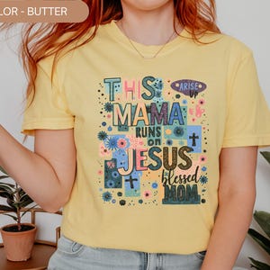 Puede incluir: Camiseta amarilla mantequilla con el texto "THIS MAMA RUNS ON JESUS blessed MOM" en un diseño floral colorido. La camiseta también tiene la palabra "ARISE" y símbolos de cruz. Un estilo casual y cómodo.