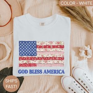 Puede incluir: Camiseta blanca con un dise&ntilde;o de la bandera estadounidense. La bandera incorpora un campo azul con estrellas blancas y secciones a rayas rojas y blancas con motivos florales. El texto "GOD BLESS AMERICA" est&aacute; bordado debajo.