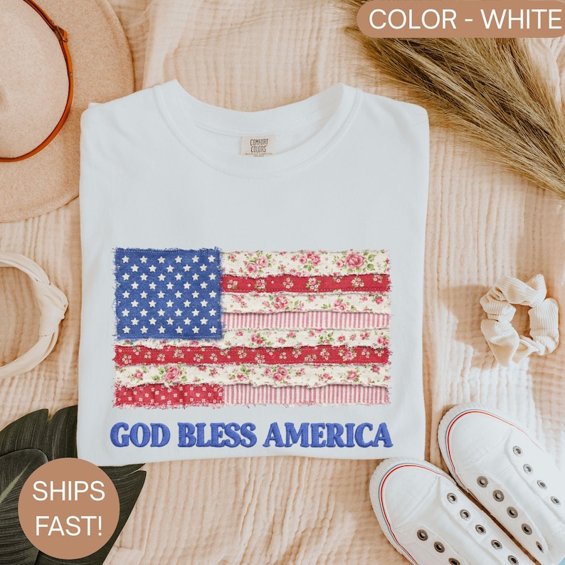 Puede incluir: Camiseta blanca con un dise&ntilde;o de la bandera estadounidense. La bandera incorpora un campo azul con estrellas blancas y secciones a rayas rojas y blancas con motivos florales. El texto "GOD BLESS AMERICA" est&aacute; bordado debajo.