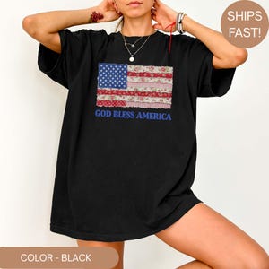 Puede incluir: Camiseta negra con un dise&ntilde;o de bandera americana de retazos. La bandera incorpora un campo azul de estrellas, rayas rojas y blancas y patrones florales. La frase "GOD BLESS AMERICA" est&aacute; impresa en azul debajo.