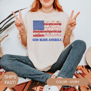 Puede incluir: Camiseta de color marfil con un dise&ntilde;o de bandera americana que incorpora patrones florales y el texto "GOD BLESS AMERICA" en azul. La camiseta es de estilo casual, adecuada para el uso diario.