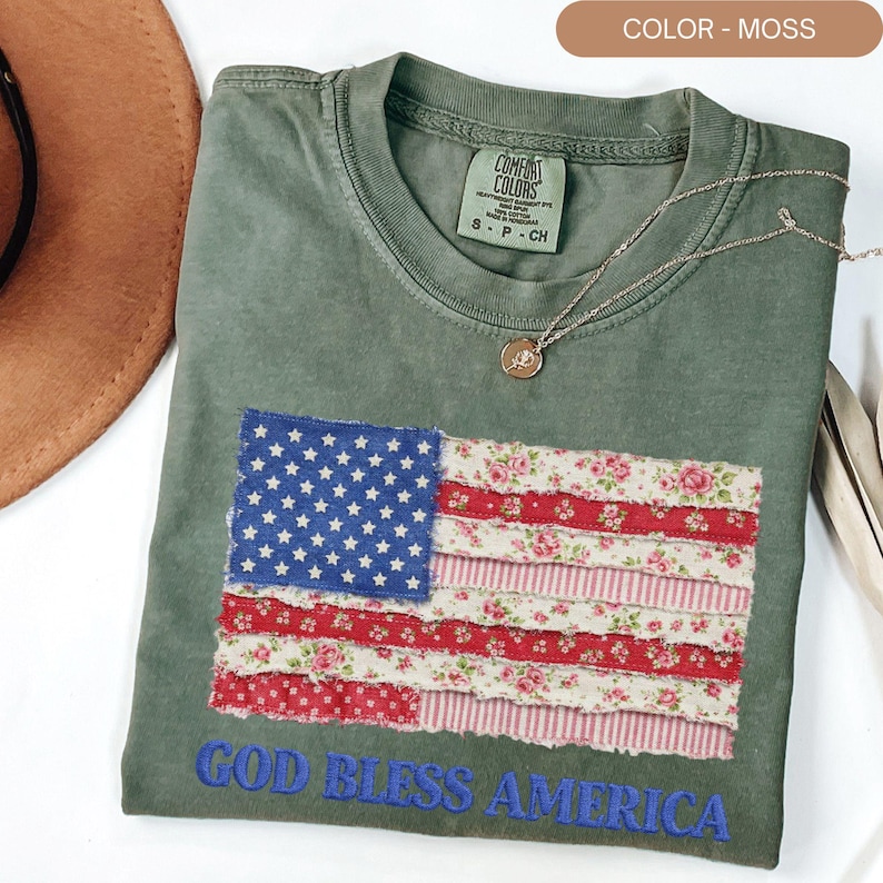 Puede incluir: Una camiseta de color musgo con un dise&ntilde;o de la bandera estadounidense. La bandera presenta un cuadrado azul con estrellas blancas y rayas rojas y florales. Las palabras "GOD BLESS AMERICA" est&aacute;n bordadas en azul.