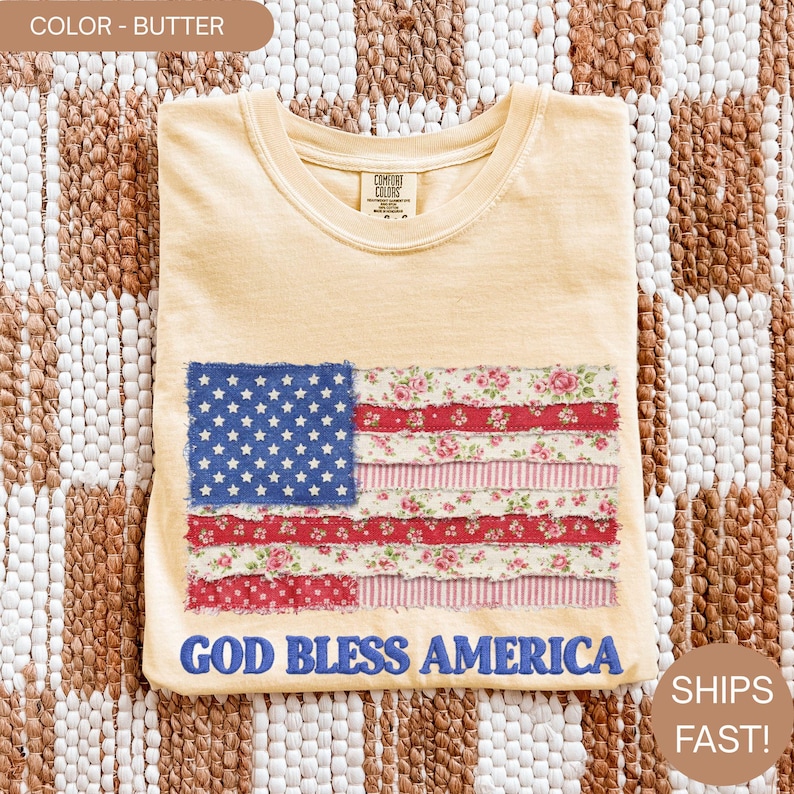 Puede incluir: Camiseta color mantequilla con un dise&ntilde;o de bandera americana en patchwork. La bandera tiene una secci&oacute;n azul con estrellas blancas y rayas rojas y florales. El texto "GOD BLESS AMERICA" est&aacute; debajo.