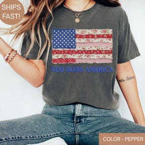 Puede incluir: Camiseta gris con un dise&ntilde;o de bandera americana de retazos con patrones florales y de estrellas. La frase "GOD BLESS AMERICA" est&aacute; bordada debajo. La camiseta es de color pimienta.