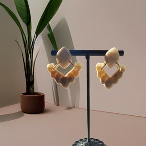 May include: A pair of beige statement earrings with a gold accent. The earrings are displayed on a black stand with a decorative base.