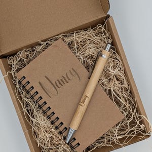 May include: A gift set in a cardboard box, featuring a spiral-bound notebook and a pen. The notebook is brown with the name "Nancy" printed on the cover. The pen is made of bamboo and also has "Nancy" printed on it. The box is filled with straw.