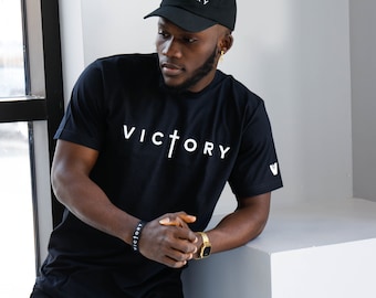 May include: Black t-shirt and baseball cap with the word "VICTORY" in white. The t-shirt has a cross design within the word. A black bracelet with the word "VICTORY" is also visible. The cap has the word "VICTORY" in white.