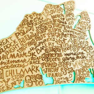 May include: A wooden map of Martha's Vineyard with the names of towns and landmarks written on it. The map is painted in a light blue color and has a white border. The names of the towns and landmarks are written in a brown color.