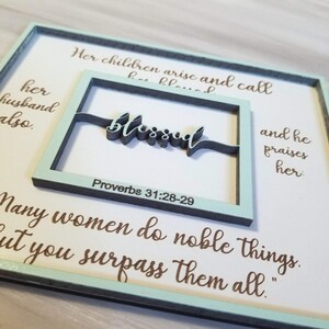 May include: A light blue wooden sign with a white background and brown text. The sign features a cutout frame with the word "blessed" in black lettering. The text "Proverbs 31:28-29" is printed below the word "blessed". The sign also includes the text "Her children arise and call her husband also. And he praises her. Many women do noble things, but you surpass them all."