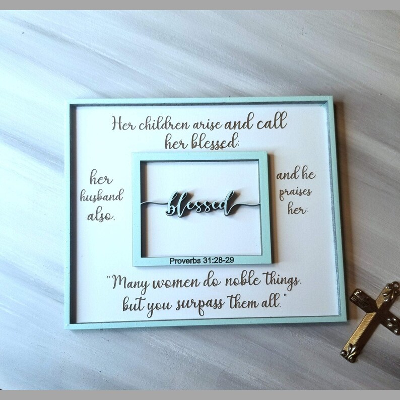 May include: A light blue framed wall art with a white background and the text "Her children arise and call her blessed: her husband also. blessed Proverbs 31:28-29 and he praises her "Many women do noble things. but you surpass them all."  A gold cross is in the bottom right corner of the image.