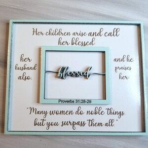 May include: A framed wood sign with a light blue border and white background. The sign features a quote from Proverbs 31:28-29, "Her children arise and call her blessed: her husband also.  "Many women do noble things, but you surpass them all." The word "blessed" is written in cursive and is a separate piece of wood.