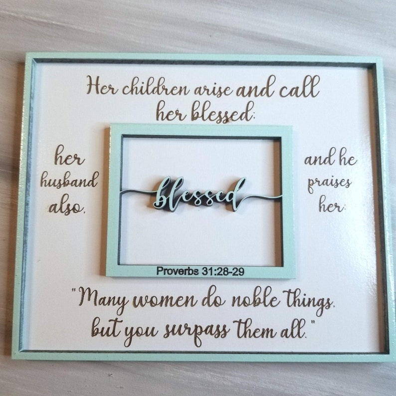 May include: A framed wood sign with a light blue border and white background. The sign features a quote from Proverbs 31:28-29, "Her children arise and call her blessed: her husband also.  "Many women do noble things, but you surpass them all." The word "blessed" is written in cursive and is a separate piece of wood.
