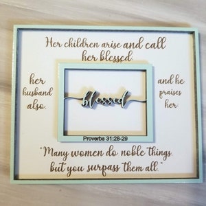 May include: A framed wood sign with a light blue background and brown text. The sign features a quote from Proverbs 31:28-29, "Her children arise and call her blessed: her husband also.  "Many women do noble things, but you surpass them all."