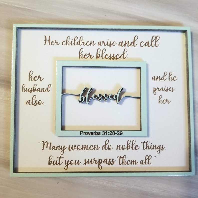 May include: A framed wood sign with a light blue background and brown text. The sign features a quote from Proverbs 31:28-29, "Her children arise and call her blessed: her husband also.  "Many women do noble things, but you surpass them all."