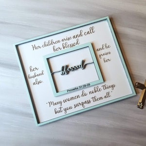 May include: A light blue wooden sign with a white background and the text "Her children arise and call her blessed. Her husband also. Blessed. Proverbs 31:28-29 "Many women do noble things, but you surpass them all."