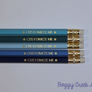 May include: Five pencils in varying shades of blue, each with a gold-coloured ferrule and eraser. The pencils are printed with the text "CUSTOMIZE ME" and a star symbol in gold. The pencils are arranged horizontally.