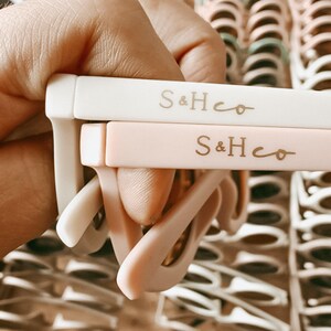 Pu&ograve; includere: Due paia di occhiali da sole con montature bianche e rosa. Le montature sono etichettate con il testo "S&Hco" in lettere dorate.