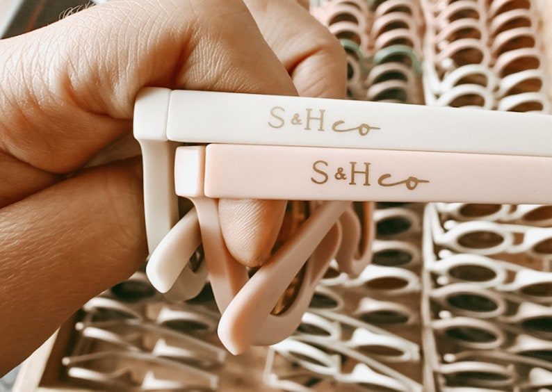 Pu&ograve; includere: Due paia di occhiali da sole con montature bianche e rosa. Le montature sono etichettate con il testo "S&Hco" in lettere dorate.