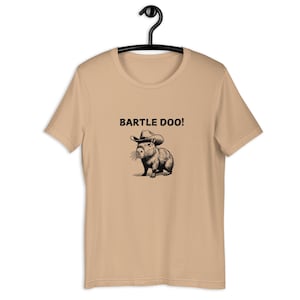 Op de afbeelding: Een beige T-shirt met een zwart-witte illustratie van een capibara die een cowboyhoed draagt. De tekst "BARTLE DOO!" staat boven de capibara.