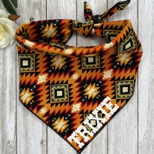 May include: A triangular pet bandana featuring a geometric pattern in shades of orange, black, cream, and teal. The fabric is tied at the top corner and displays the name TRIXIE in bold white capital letters across the bottom right corner.